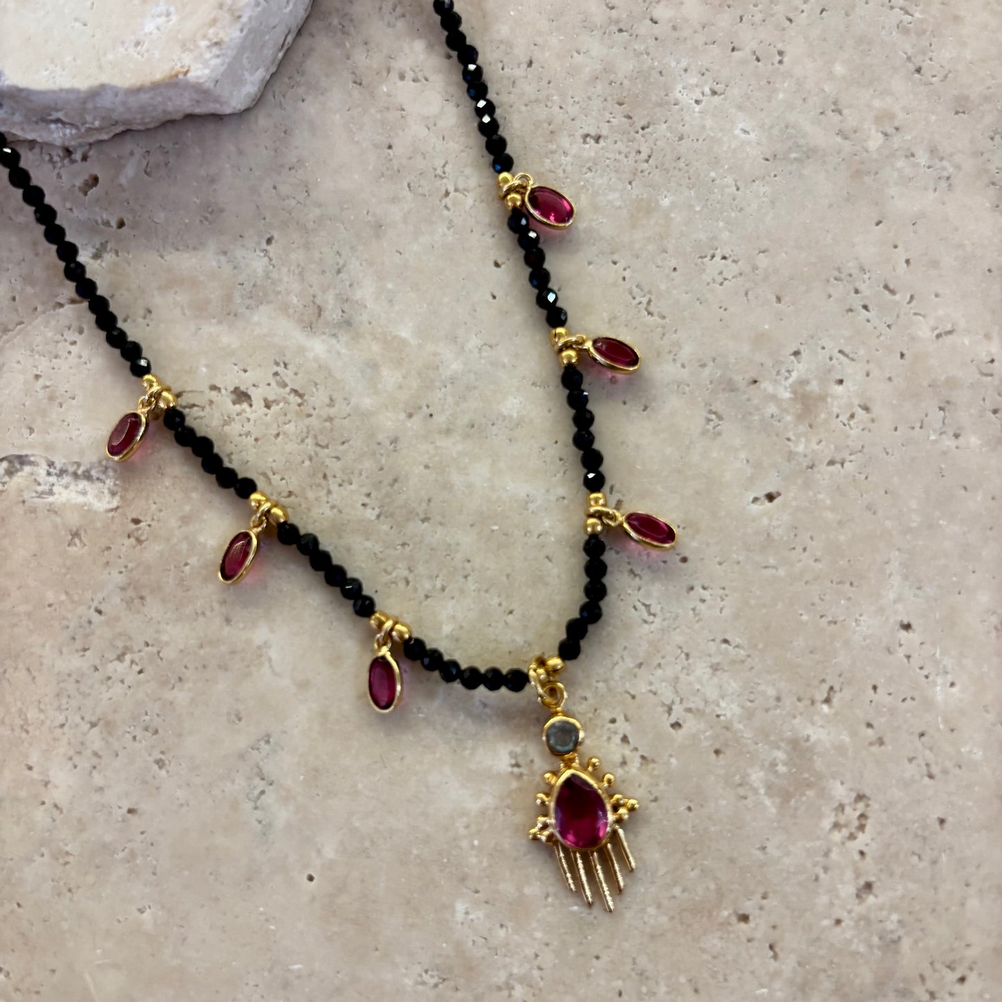Collier Birdy noir composé de perles d’onyx noir et de pampilles en quartz framboise serties or, avec pendentif central assorti, présenté sur une surface minérale beige.