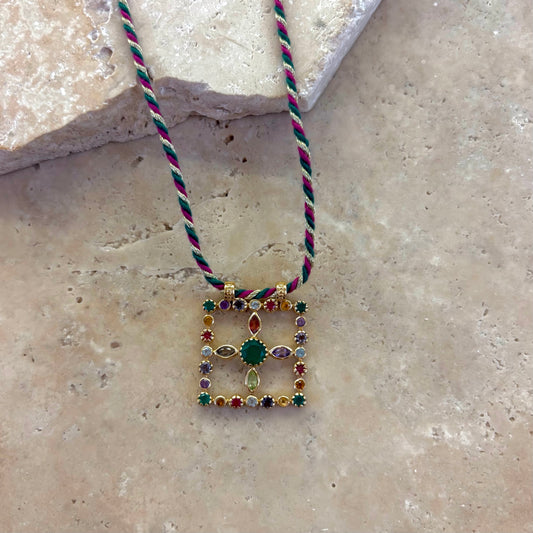 Collier Pondi onyx vert avec pendentif carré serti de pierres fines multicolores et d’un onyx vert central, monté sur un cordon torsadé vert, rose et noir, présenté sur une surface en pierre claire.