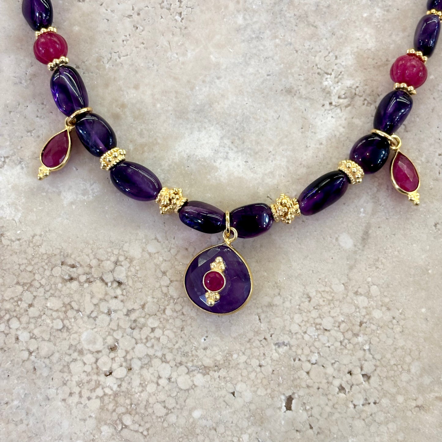Détail du collier Antonia en améthyste et quartz framboise, avec intercalaires plaqué or et pendentif rond en améthyste orné d’un cabochon framboise, présenté sur une surface en pierre claire.