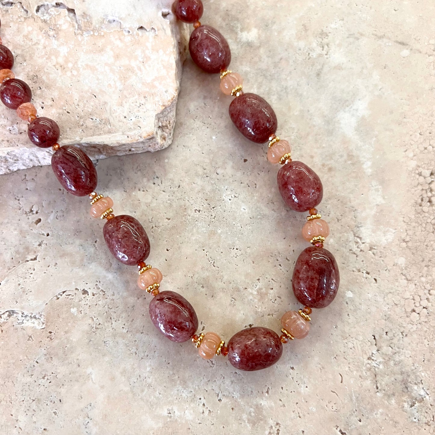 Détail du collier Gaby en quartz framboise et pierre de lune pêche, avec intercalaires dorés, présenté sur une surface minérale beige.