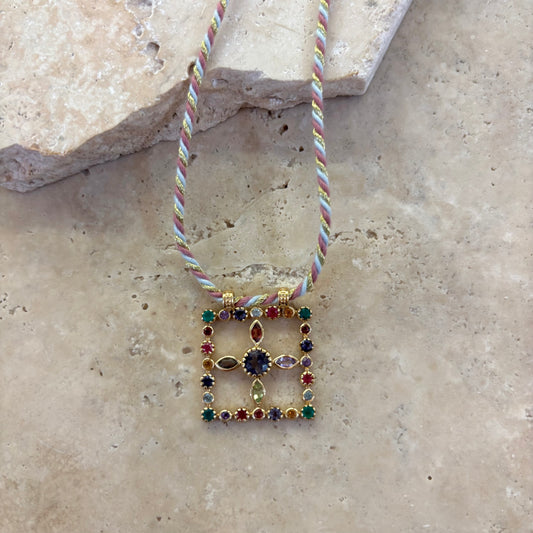 Collier Pondi améthyste avec pendentif carré en pierres fines multicolores serties or, rehaussé d’une améthyste centrale, suspendu à un cordon torsadé pastel, présenté sur une surface en pierre claire.