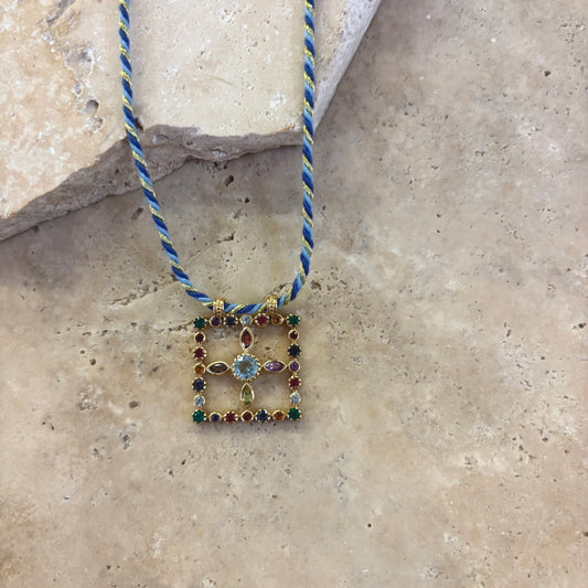 Collier Pondi topaze bleue avec pendentif carré en pierres fines multicolores serties or, orné d’une topaze bleue centrale, suspendu à un cordon torsadé bleu et or, présenté sur une surface en pierre claire.