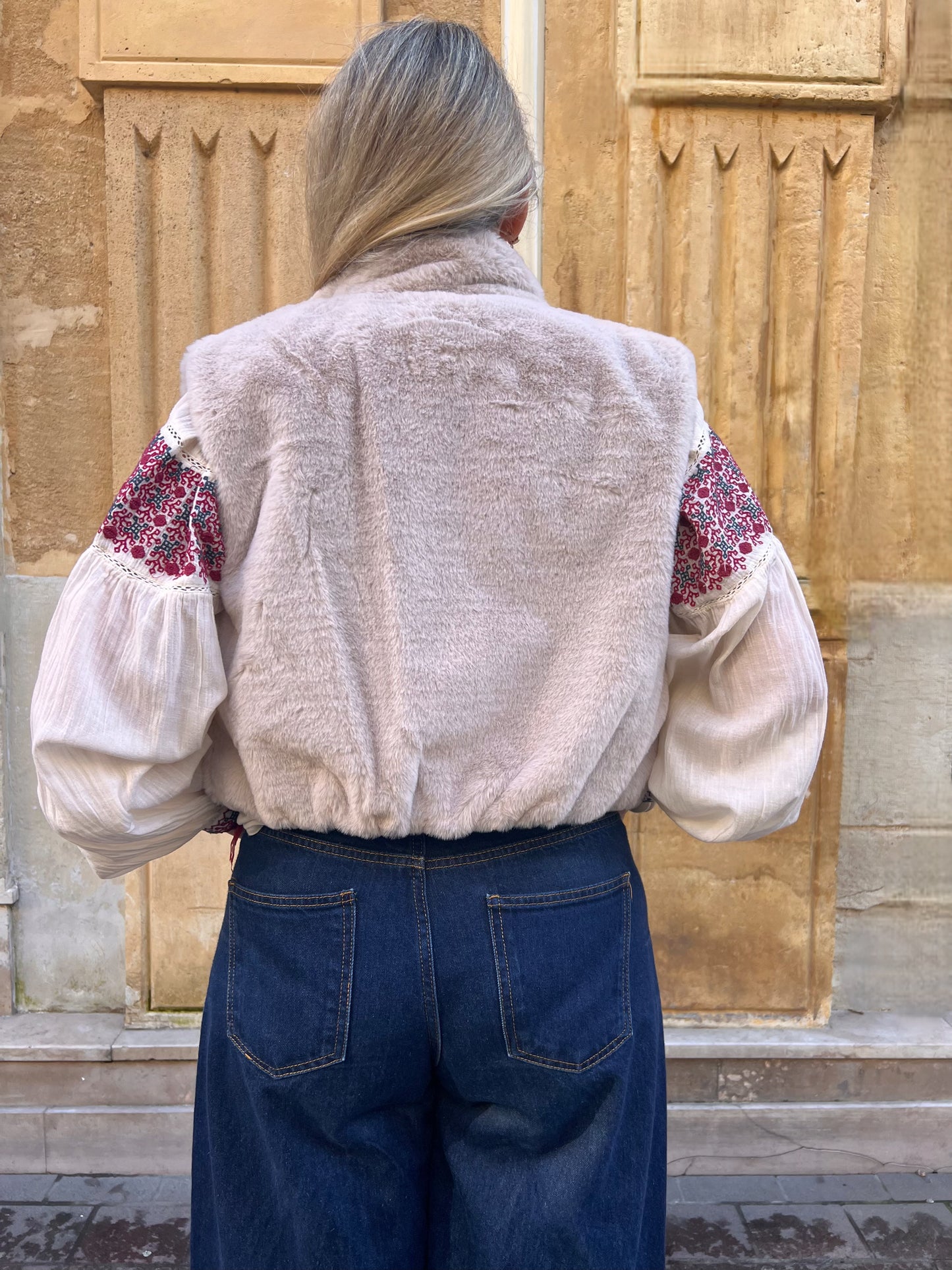 Gilet sans manches beige en fausse fourrure, porté ouvert sur une blouse imprimée rouge et un jean taille haute, vue de dos