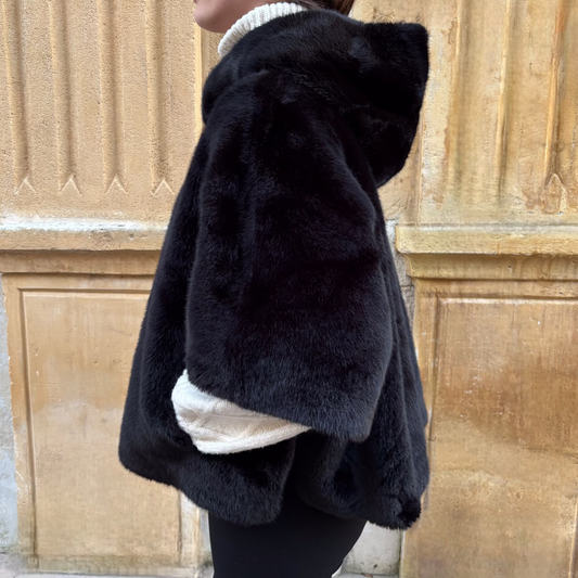 Cape noire en fausse fourrure, portée de face, fermeture zippée, coupe ample avec manches courtes et pull torsadé écru en dessous