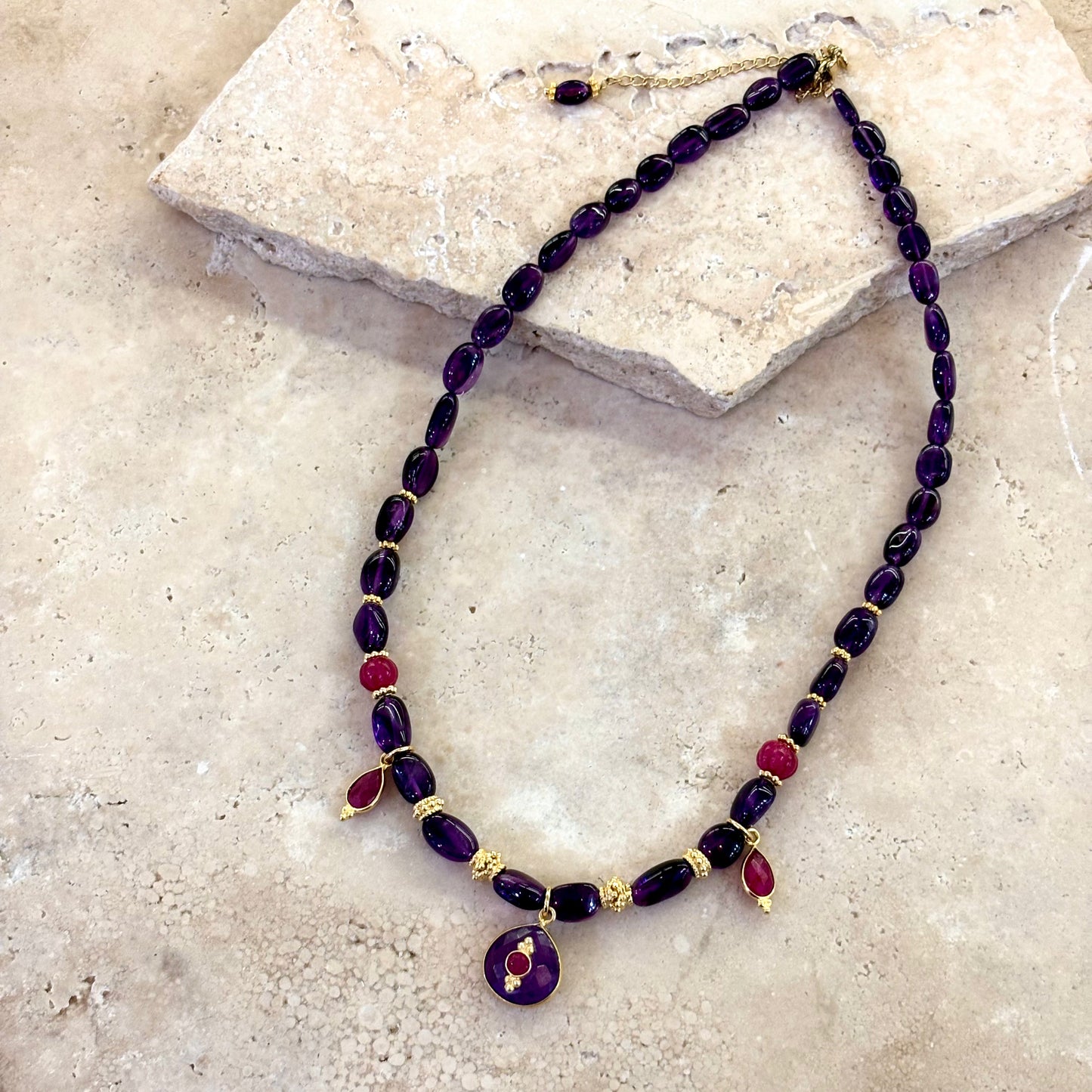 Collier Antonia composé d’améthystes profondes et de quartz framboise, avec intercalaires plaqué or et pendentif central en améthyste surmonté d’un cabochon framboise, présenté sur une surface en pierre claire.
