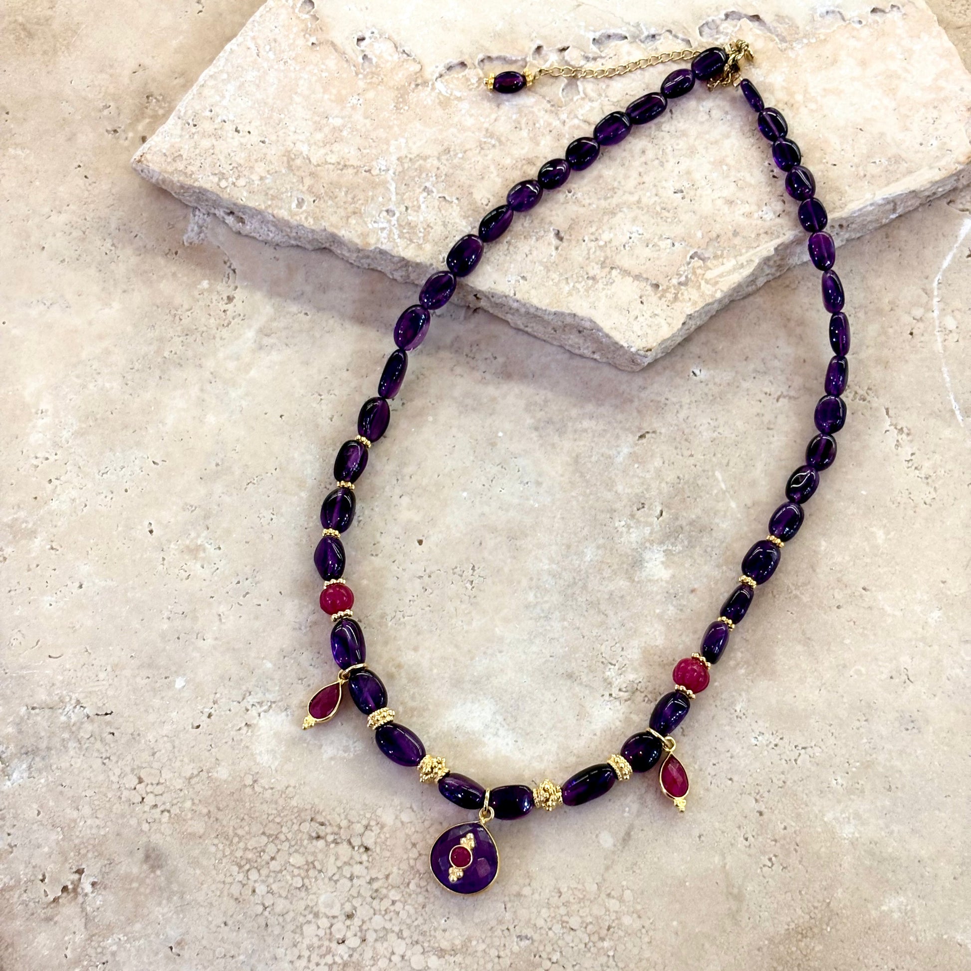 Collier Antonia composé d’améthystes profondes et de quartz framboise, avec intercalaires plaqué or et pendentif central en améthyste surmonté d’un cabochon framboise, présenté sur une surface en pierre claire.