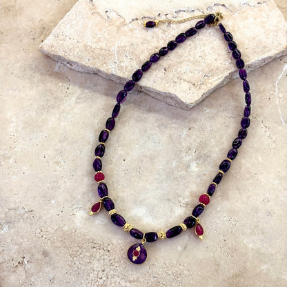 Collier Antonia composé d’améthystes profondes et de quartz framboise, avec intercalaires plaqué or et pendentif central en améthyste surmonté d’un cabochon framboise, présenté sur une surface en pierre claire.