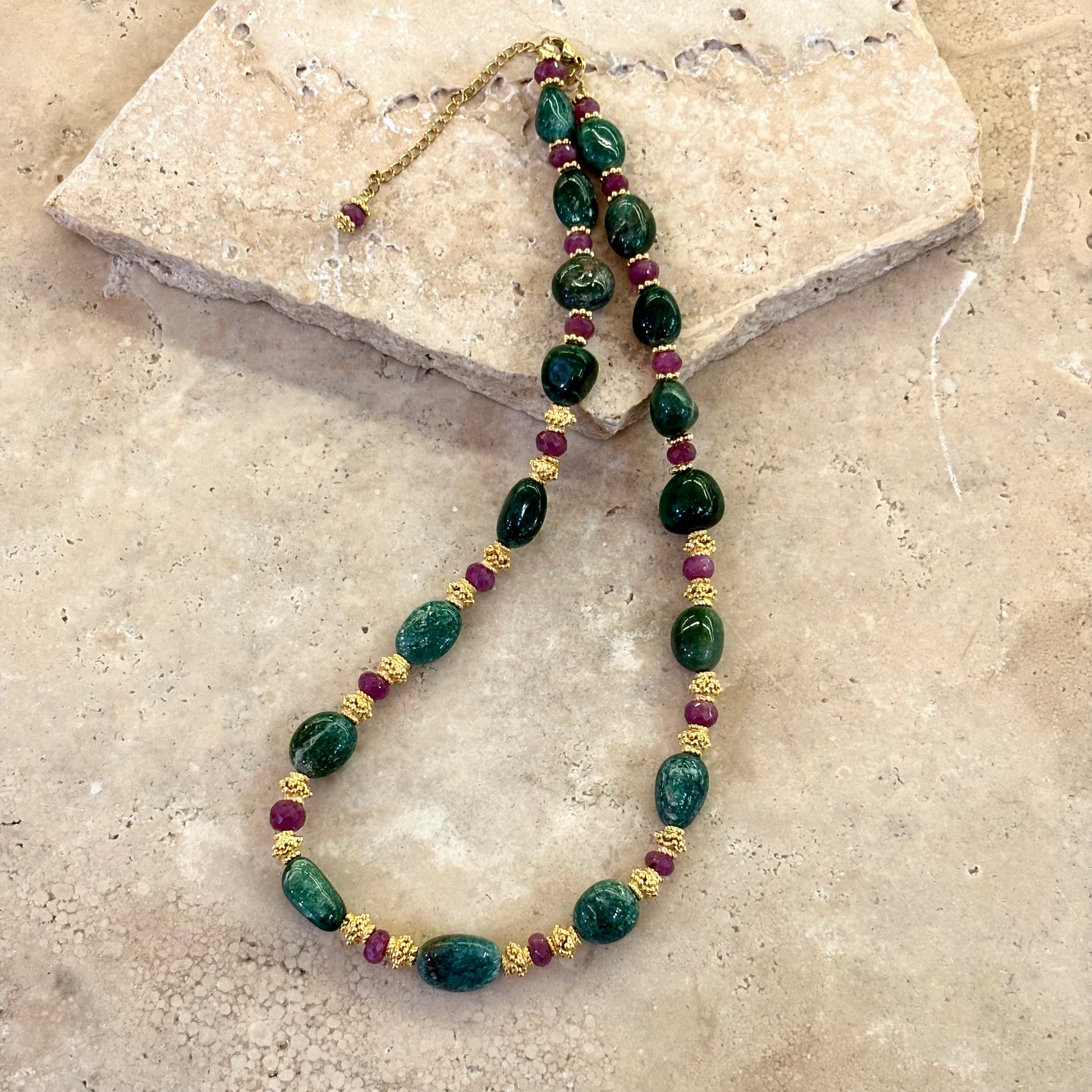 Collier Azalée composé d’aventurines vert intense et de rubis facettés, agrémenté d’intercalaires plaqués or et posé sur une pierre beige.