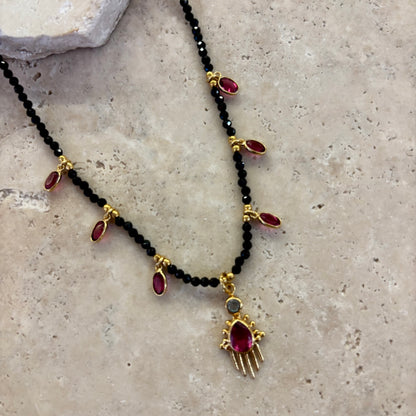 Collier Birdy noir composé de perles d’onyx noir et de pampilles en quartz framboise serties or, avec pendentif central assorti, présenté sur une surface minérale beige.