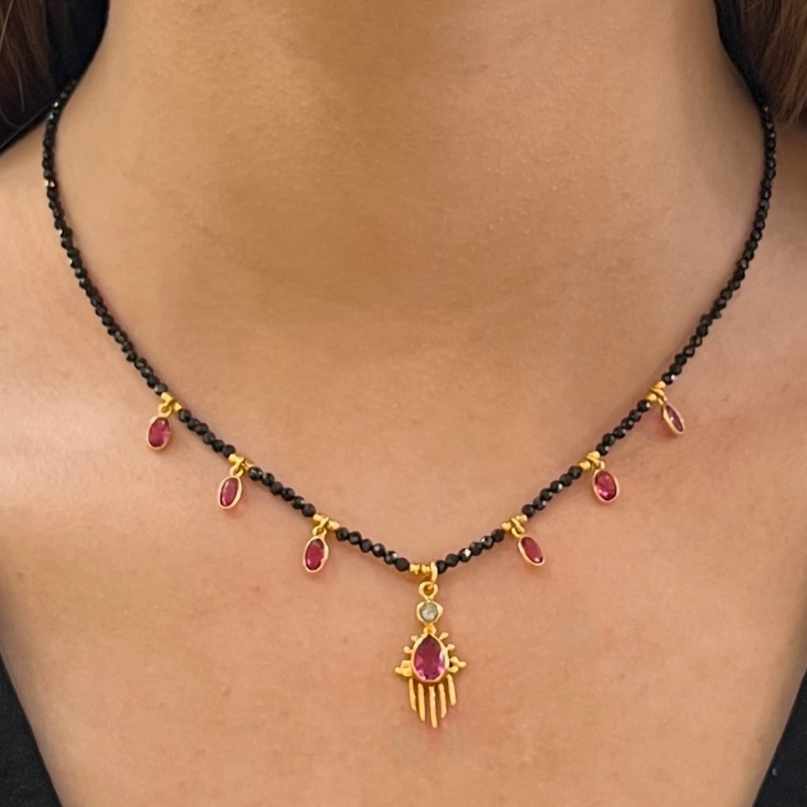 Collier Birdy porté, composé de perles d’onyx noir, de pampilles en quartz framboise et d’un pendentif central doré orné d’une pierre framboise