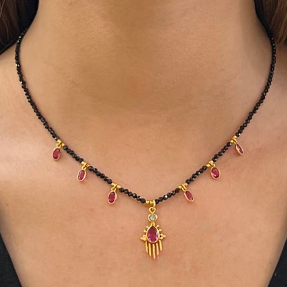Collier Birdy porté, composé de perles d’onyx noir, de pampilles en quartz framboise et d’un pendentif central doré orné d’une pierre framboise