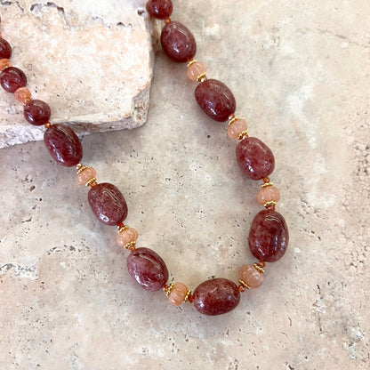 Détail du collier Gaby en quartz framboise et pierre de lune pêche, avec intercalaires dorés, présenté sur une surface minérale beige.