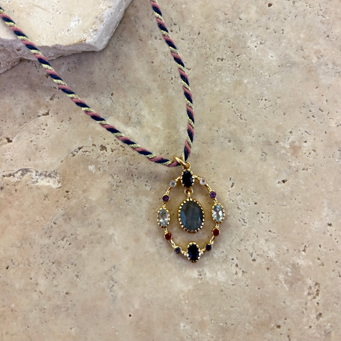 Collier Goa en labradorite avec pendentif ovale serti d’une pierre centrale grise aux reflets bleutés, entourée de pierres fines multicolores, monté sur un cordon torsadé dans les tons rose, vert et noir, présenté sur une surface en pierre claire.