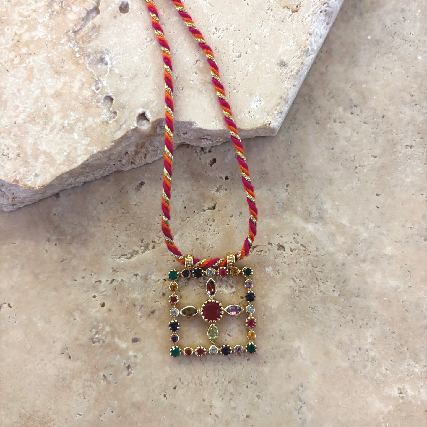 Collier Pondi avec pendentif carré en pierres fines multicolores serties or, suspendu à un cordon torsadé rouge et doré, photographié sur une surface en pierre claire.