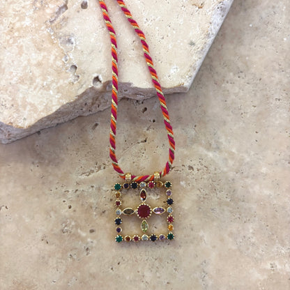 Collier Pondi avec pendentif carré en pierres fines multicolores serties or, suspendu à un cordon torsadé rouge et doré, photographié sur une surface en pierre claire.