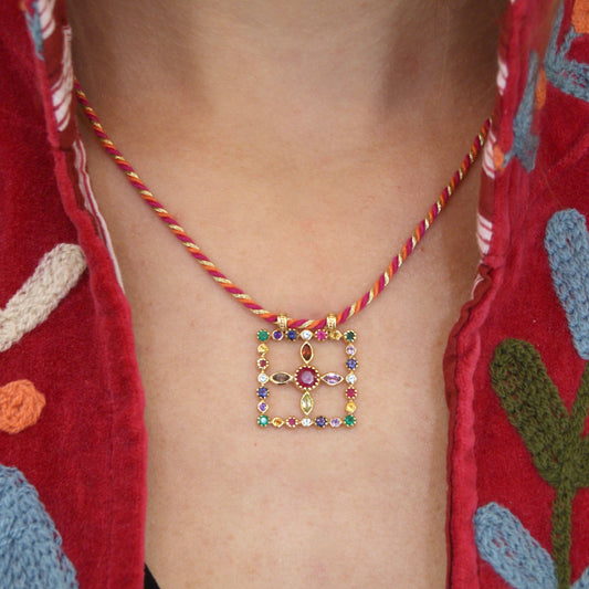 Collier Pondy porté, composé d’un pendentif carré serti de pierres fines multicolores autour d’un cabochon central framboise, suspendu à un cordon torsadé rouge et doré.