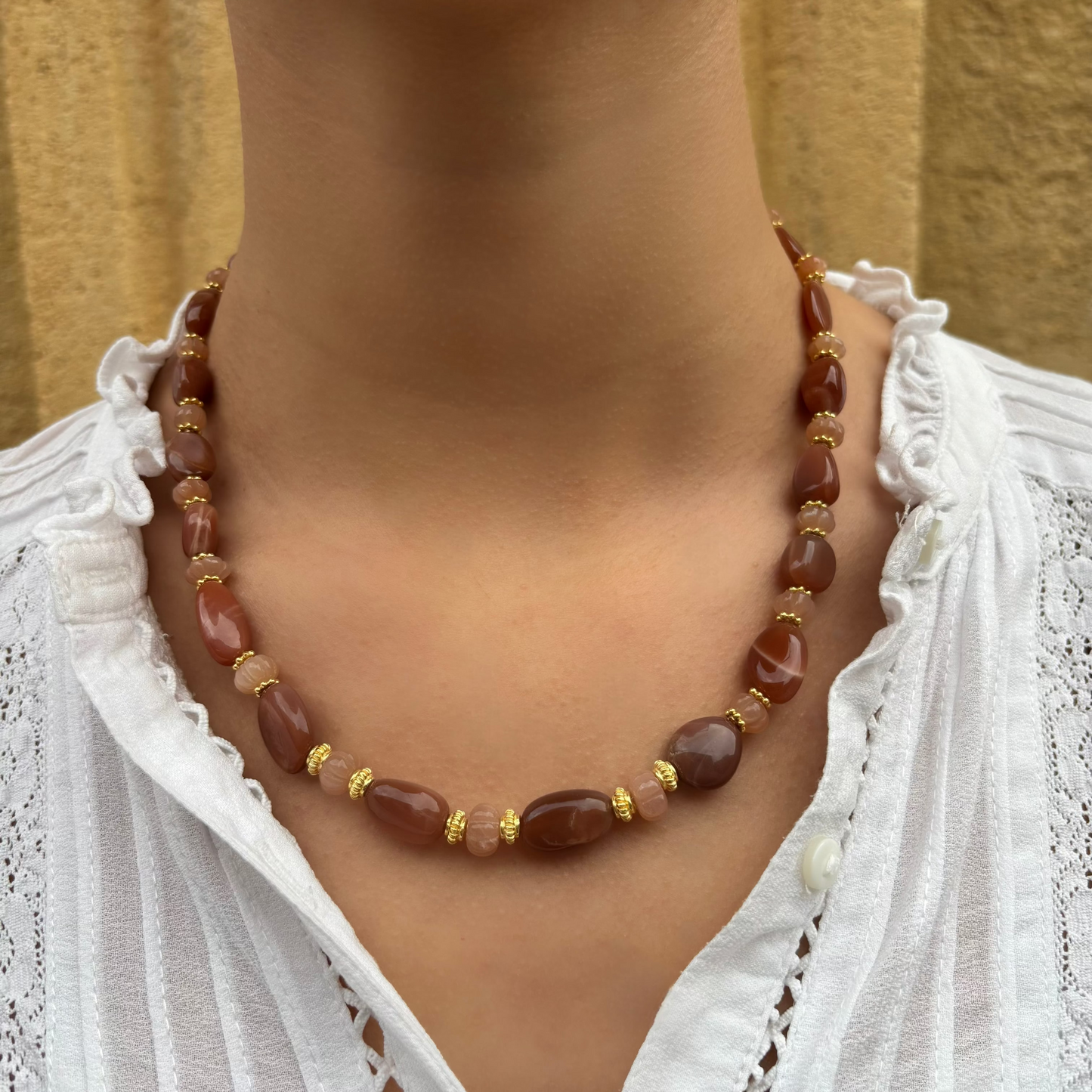 Collier Solea en pierre de lune pêche porté au cou, composé de galets polis et de petites perles sculptées en melons, rehaussé d’intercalaires plaqués or