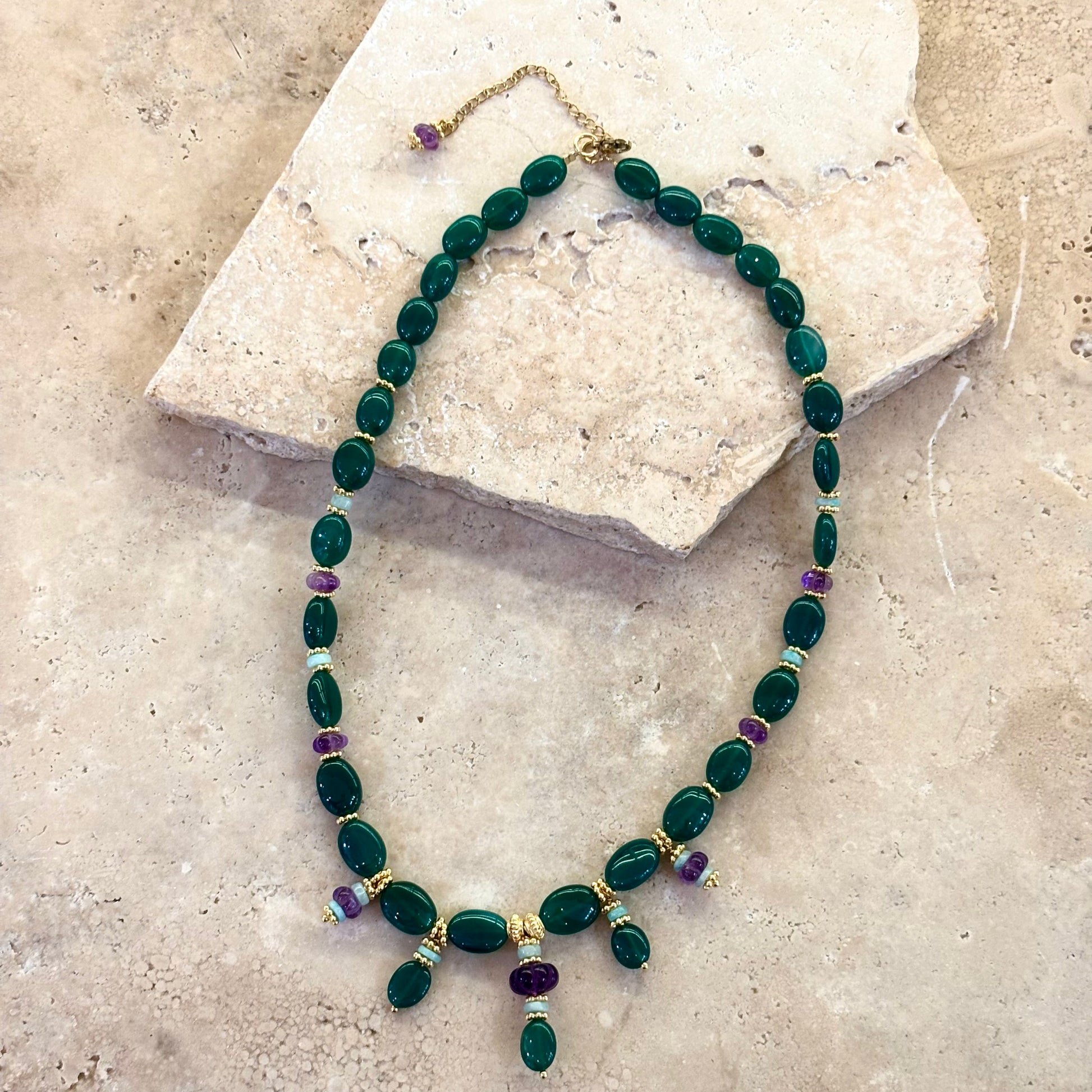 Collier Tiana composé de perles d’onyx vert et d’améthystes, agrémenté d’intercalaires plaqués or, présenté à plat sur une pierre naturelle.