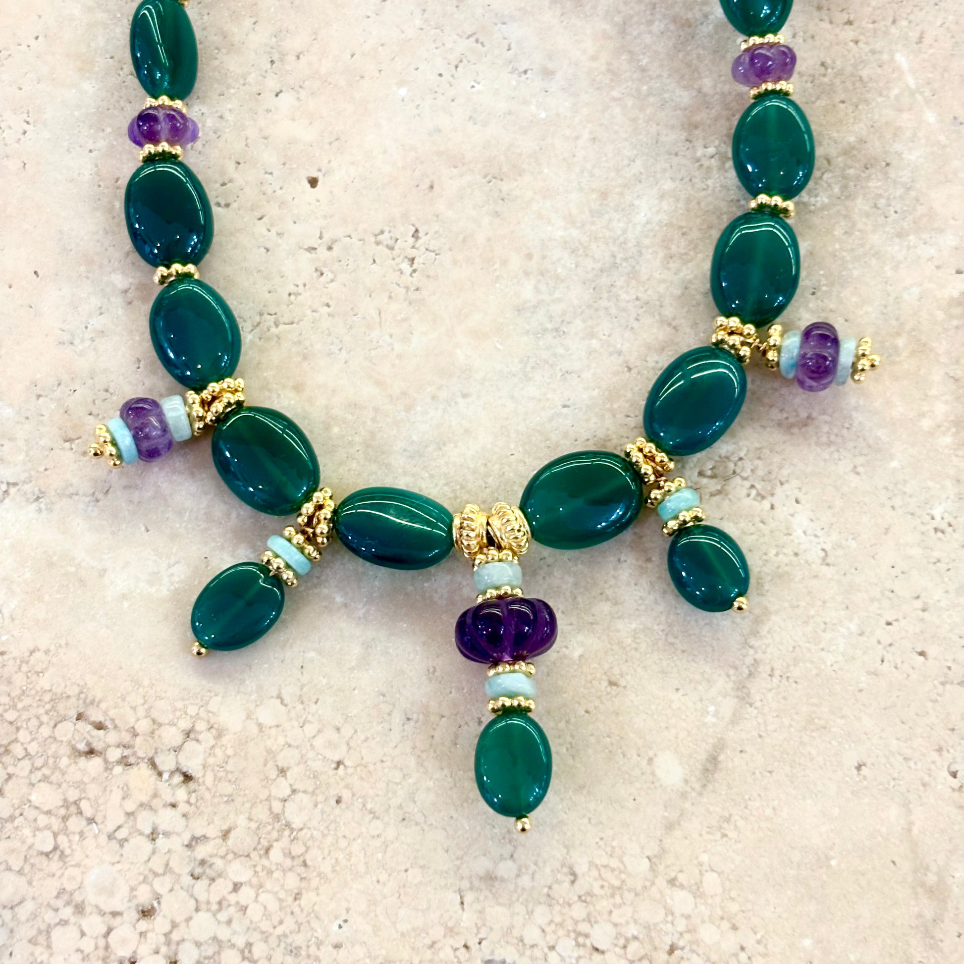 Détail du collier Tiana montrant les perles d’onyx vert, les améthystes facettées et les intercalaires plaqués or, photographiés sur une surface pierre naturelle.
