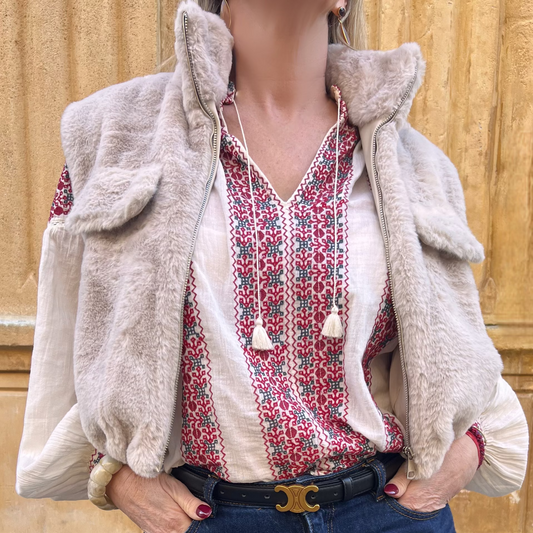 Gilet sans manches beige en fausse fourrure, porté ouvert sur une blouse imprimée rouge et un jean taille haute, vue de face