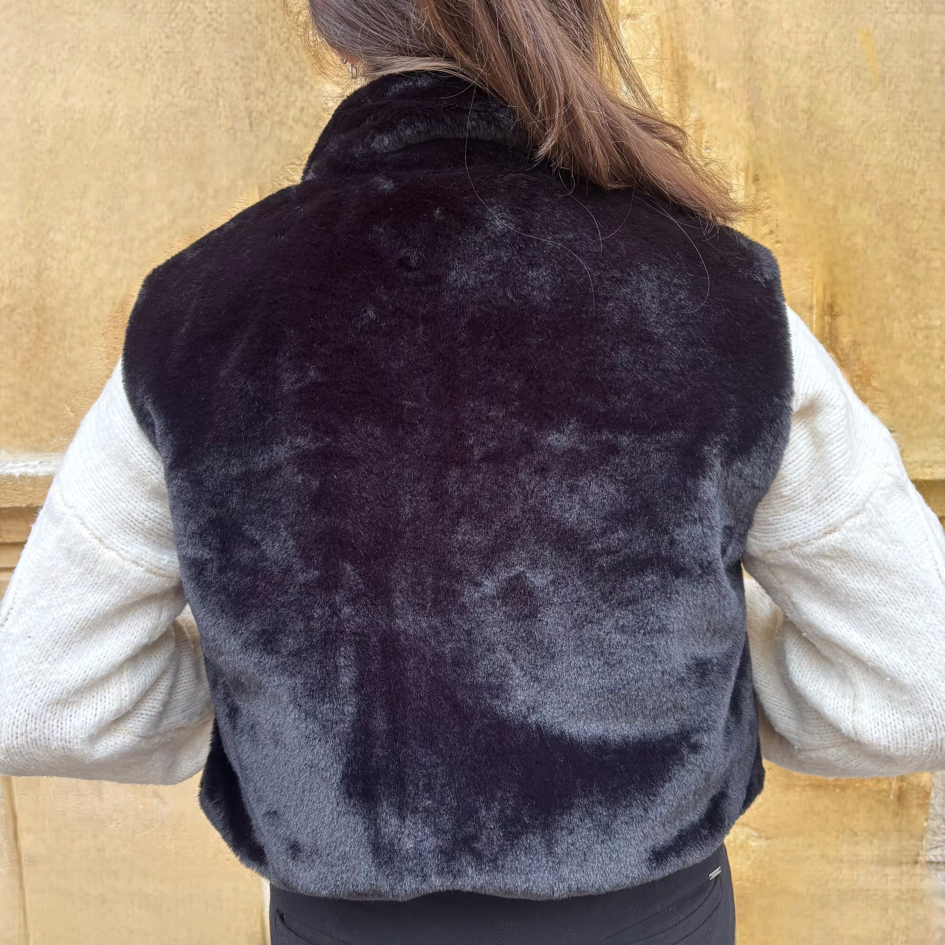 Gilet sans manches noir en fausse fourrure, porté sur un pull torsadé écru, vue de dos