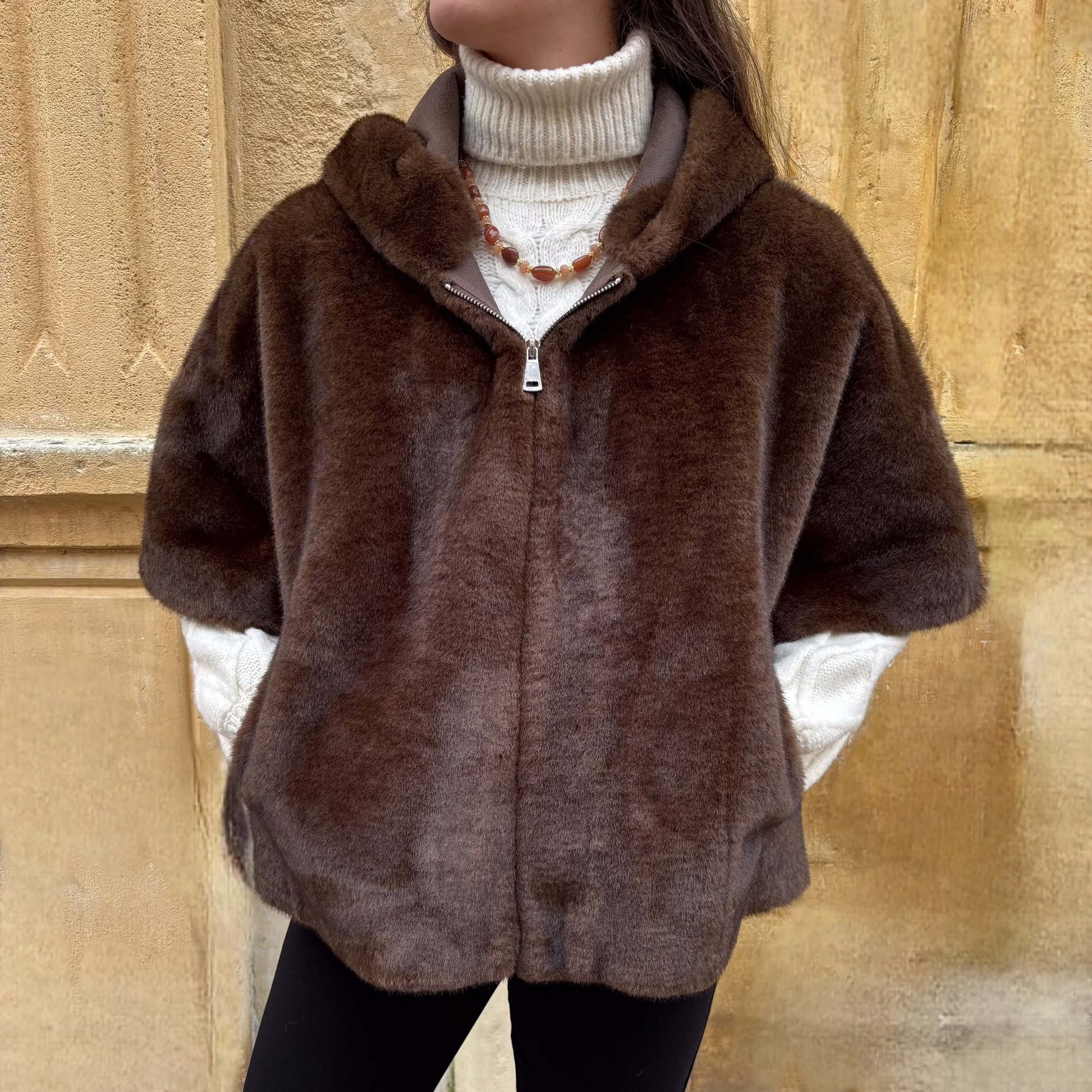 Cape marron en fausse fourrure portée de face, avec fermeture zippée et col montant, superposée à un pull torsadé écru