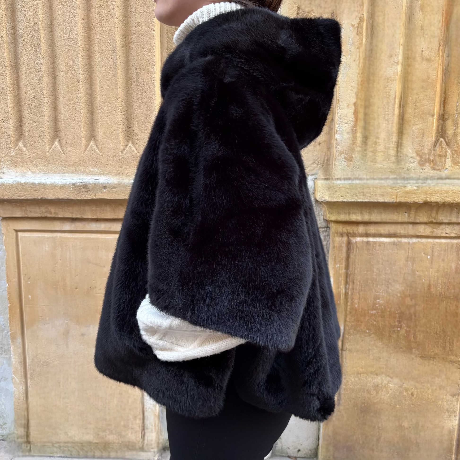 Cape noire en fausse fourrure, portée de face, fermeture zippée, coupe ample avec manches courtes et pull torsadé écru en dessous