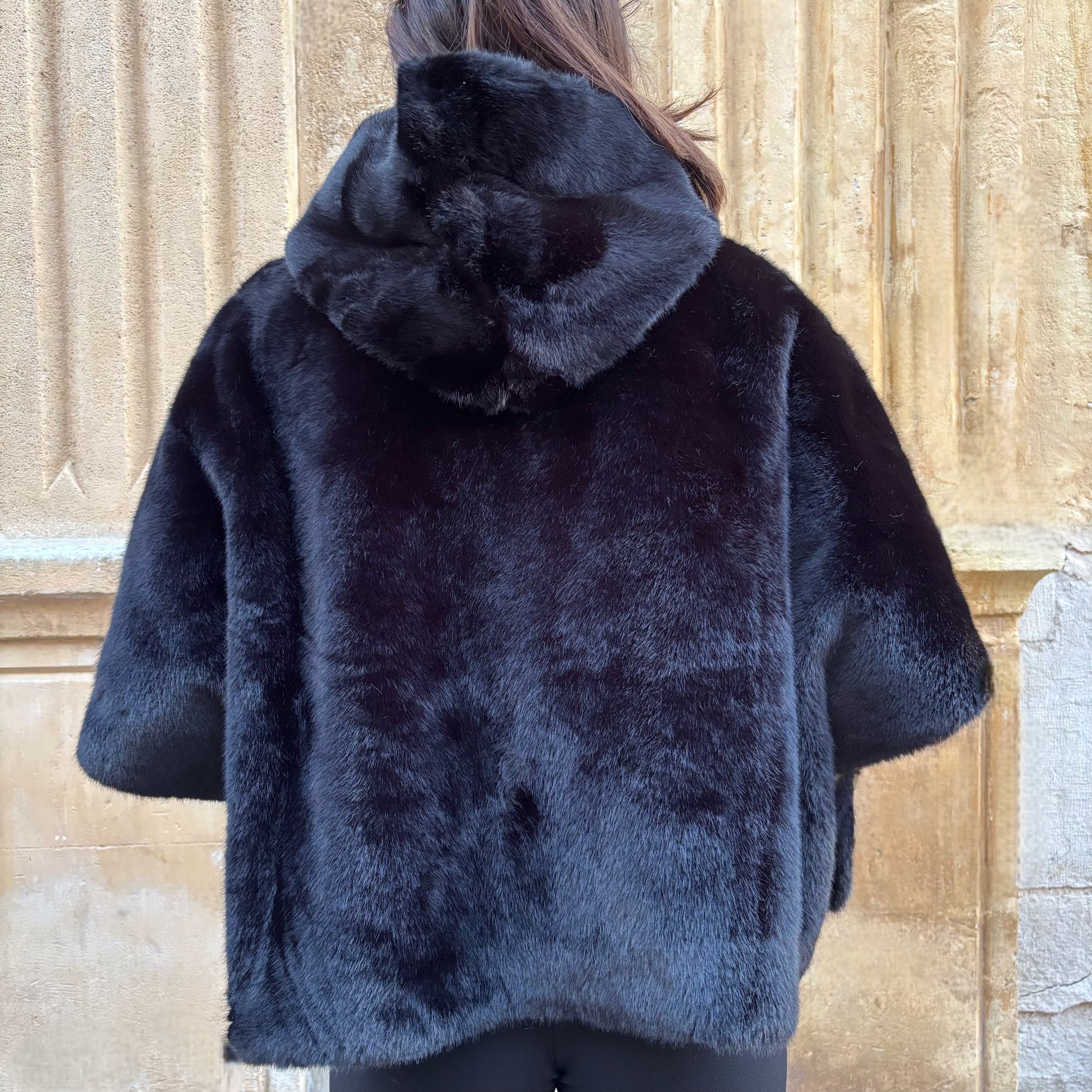 Cape noire en fausse fourrure, portée de face, fermeture zippée, coupe ample avec manches courtes et pull torsadé écru en dessous