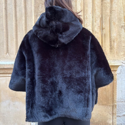 Cape noire en fausse fourrure, portée de face, fermeture zippée, coupe ample avec manches courtes et pull torsadé écru en dessous
