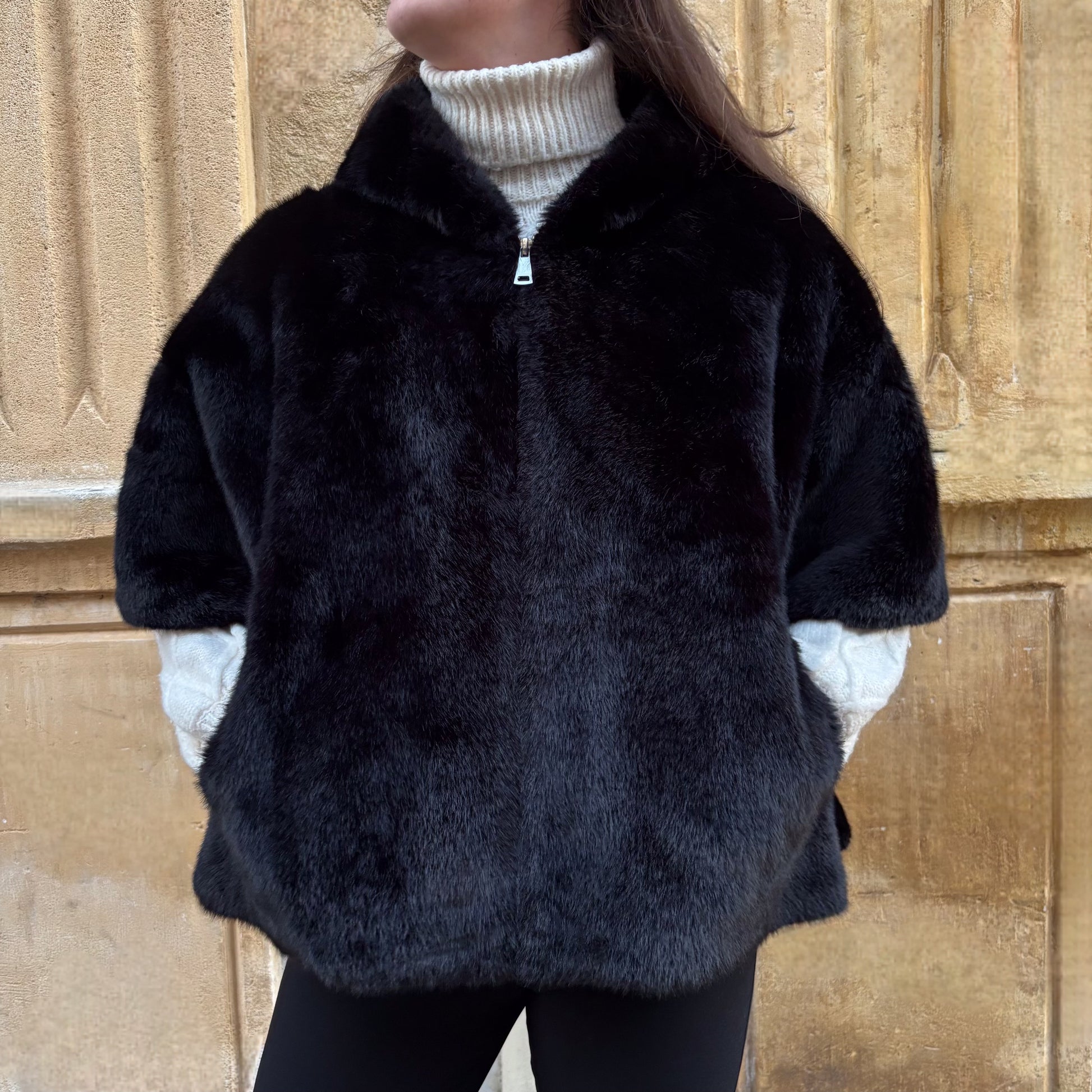 Cape noire en fausse fourrure, portée de face, fermeture zippée, coupe ample avec manches courtes et pull torsadé écru en dessous
