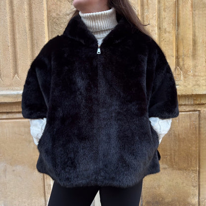 Cape noire en fausse fourrure, portée de face, fermeture zippée, coupe ample avec manches courtes et pull torsadé écru en dessous