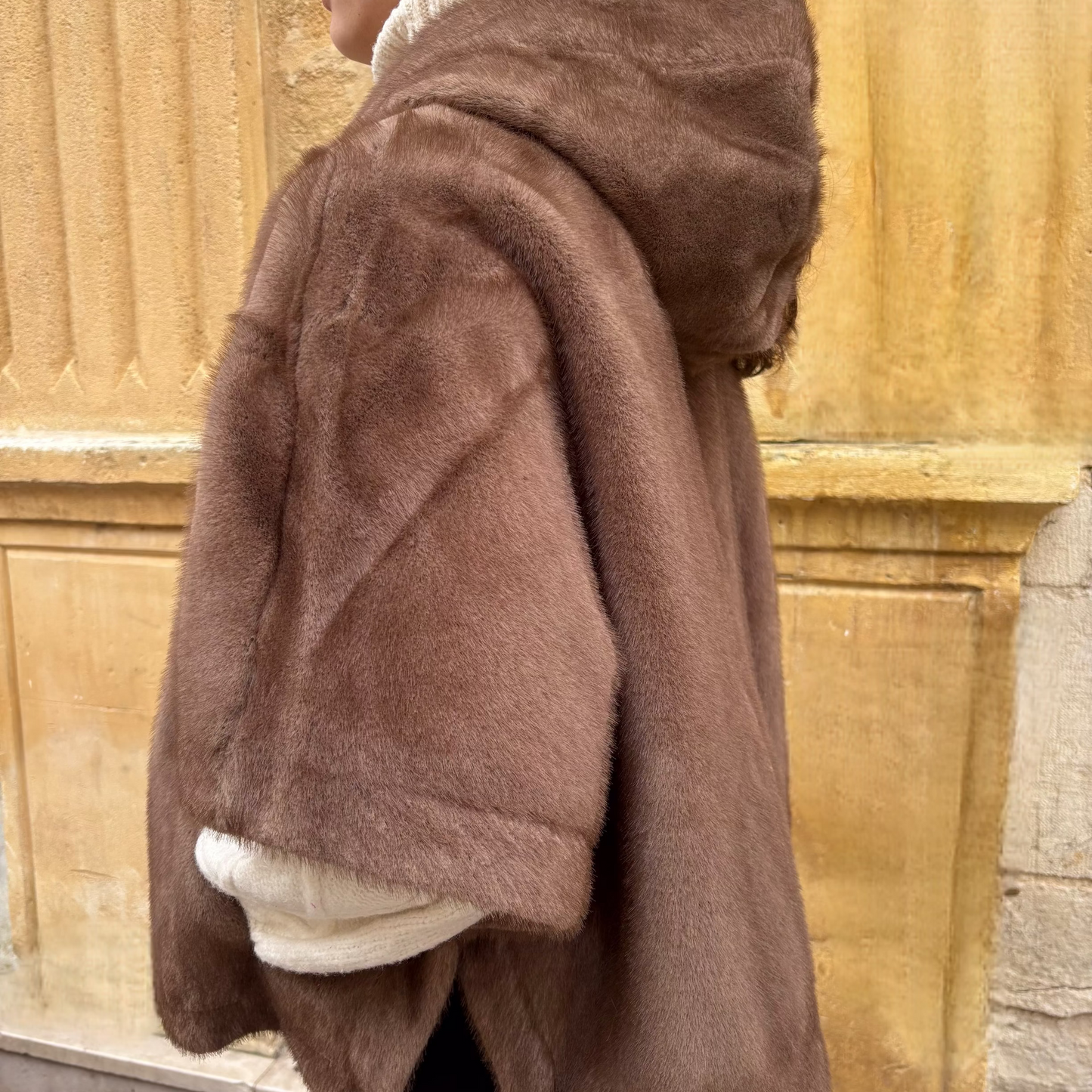 Cape taupe en fausse fourrure avec capuche, portée de face, fermeture zippée et manches courtes, superposée à un pull écru
