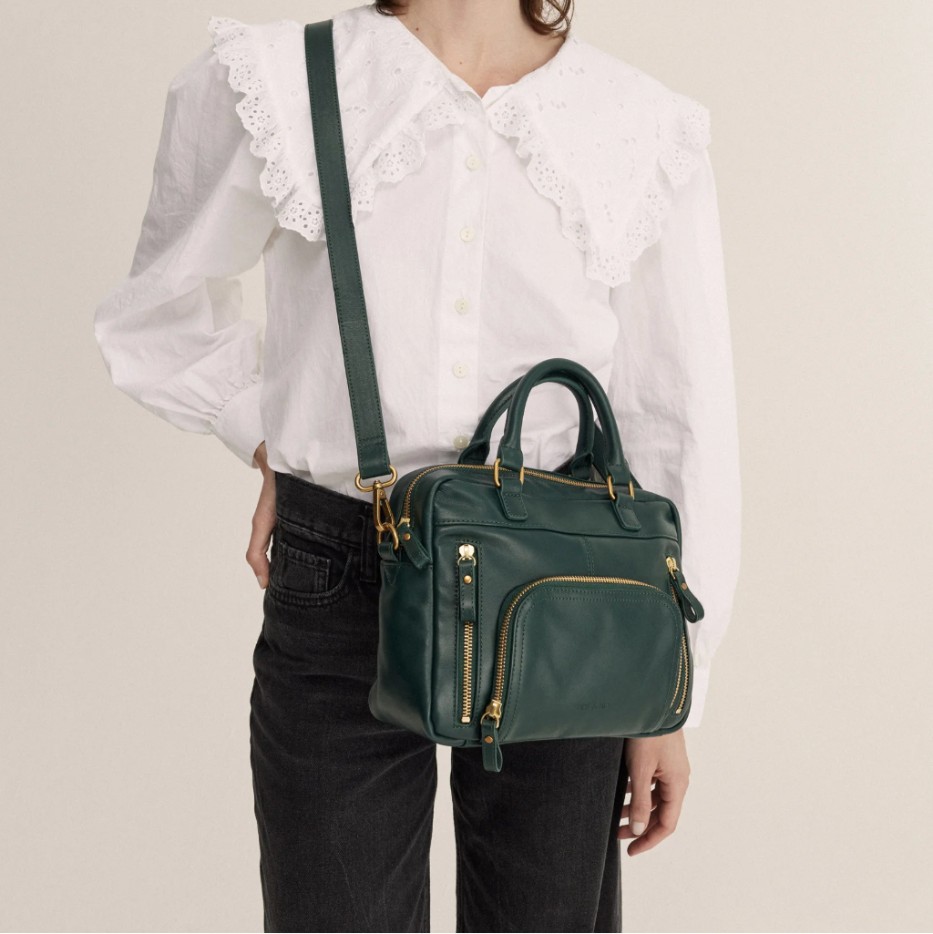 Femme portant le sac Mini Macy vert sapin Nat & Nin en bandoulière, petit sac en cuir avec poches zippées et anses renforcées.