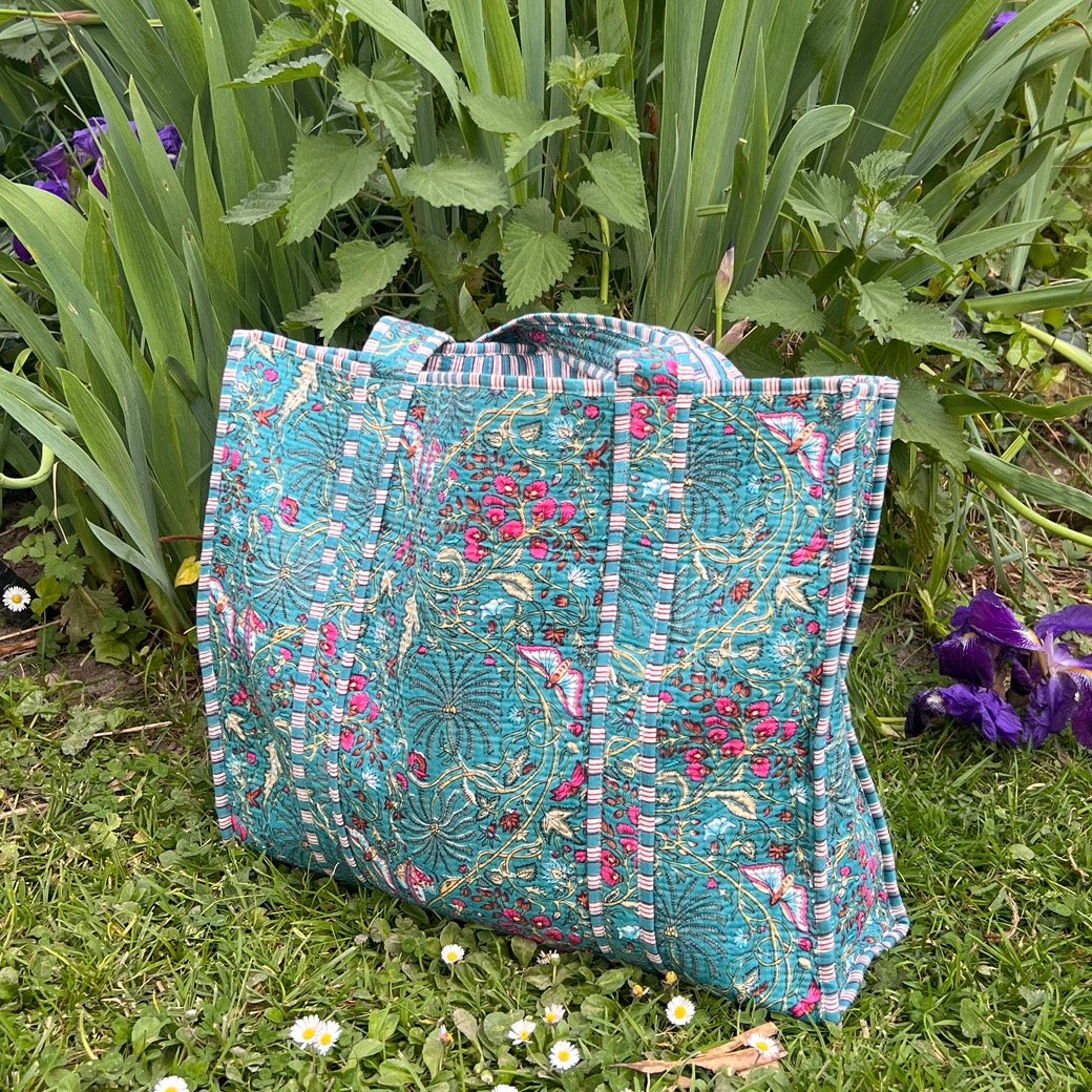 Sac cabas Butterfly en coton matelassé turquoise à motifs floraux, création artisanale Mademoiselle Azalée à Agen, posé sur l’herbe devant un massif de fleurs