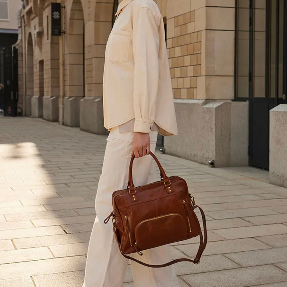 Sac Macy cognac glacé de Nat & Nin porté à la main par une femme en tenue claire, devant un bâtiment urbain.
