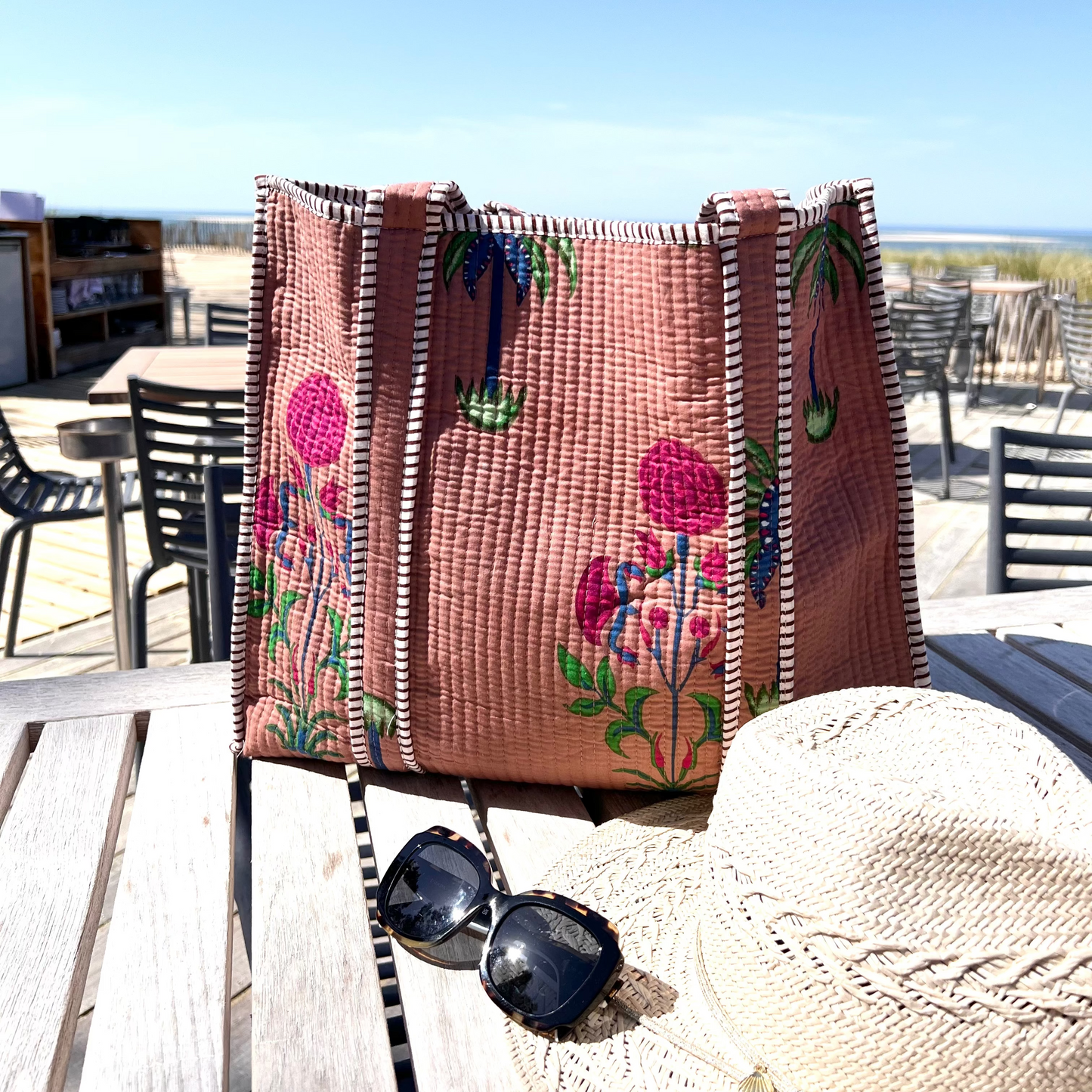 Sac cabas Palmito en coton matelassé, création Mademoiselle Azalée à Agen, posé sur une table en terrasse avec chapeau de paille et lunettes de soleil, face à une vue de plage