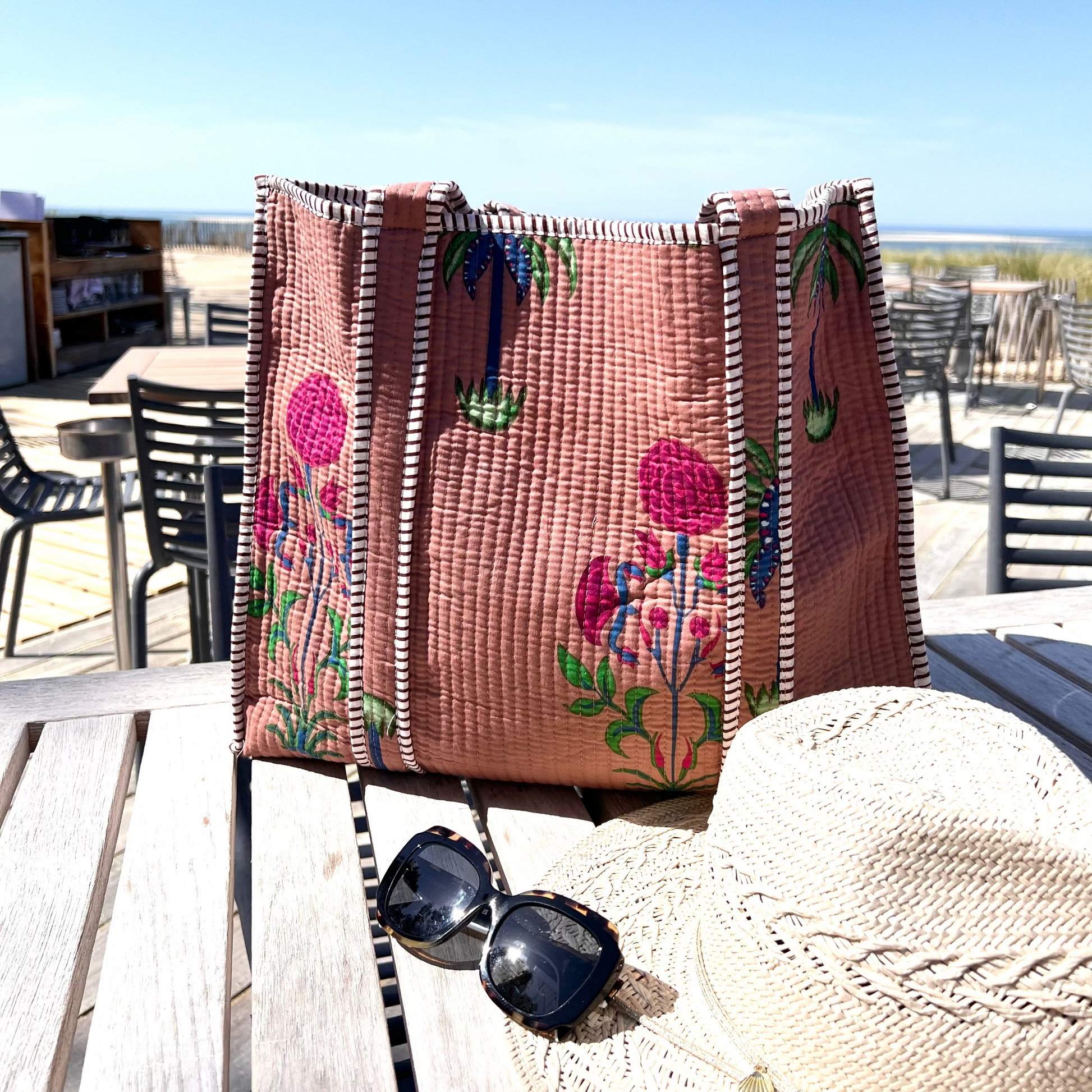 Sac cabas Palmito en coton matelassé, création Mademoiselle Azalée à Agen, posé sur une table en terrasse avec chapeau de paille et lunettes de soleil, face à une vue de plage