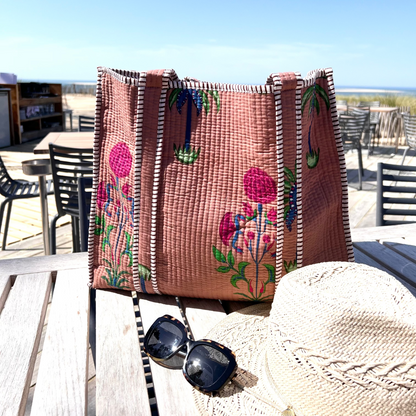 Sac cabas Palmito en coton matelassé, création Mademoiselle Azalée à Agen, posé sur une table en terrasse avec chapeau de paille et lunettes de soleil, face à une vue de plage
